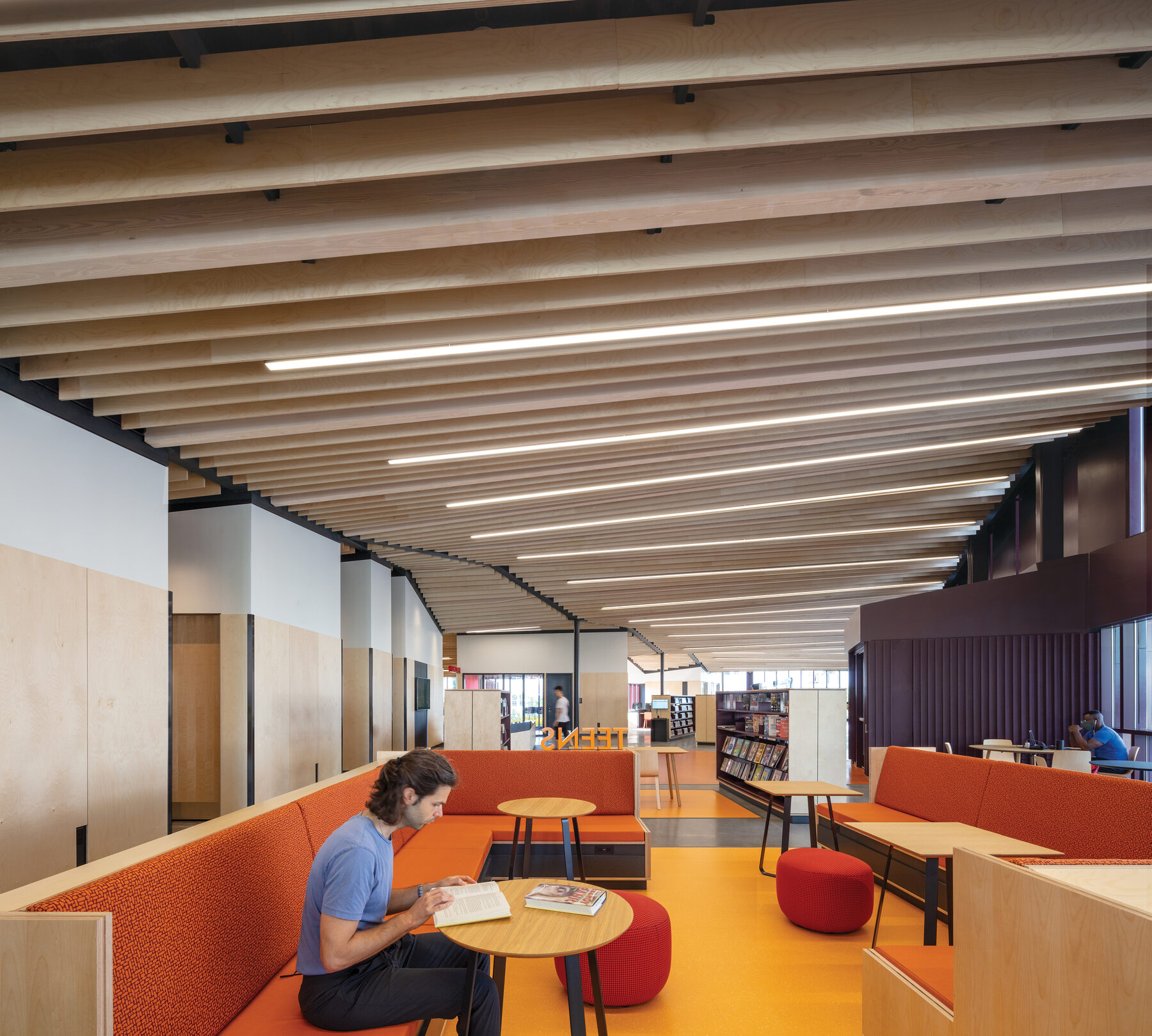 Teen wing with orange seating, a person reading at the table, and the rolling vaulted ceiling above
