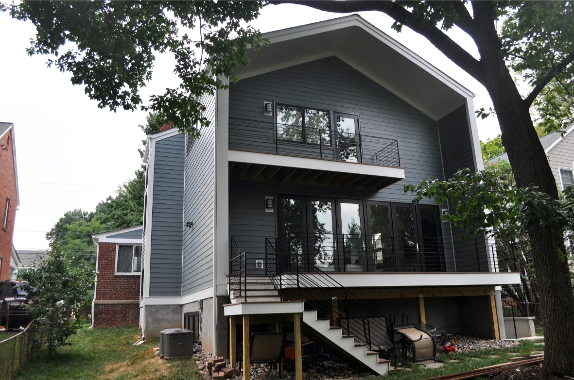 Rear elevation of the renovated house: a tall dark gray gable with deep overhanging eaves over a two-story addition, a full-height wall of black-framed windows on the main level opening onto a deck with cable rails, a smaller balcony off the upper-floor master bedroom, and the brick chimney of the original 1930s colonial visible at left between the addition and the neighbor