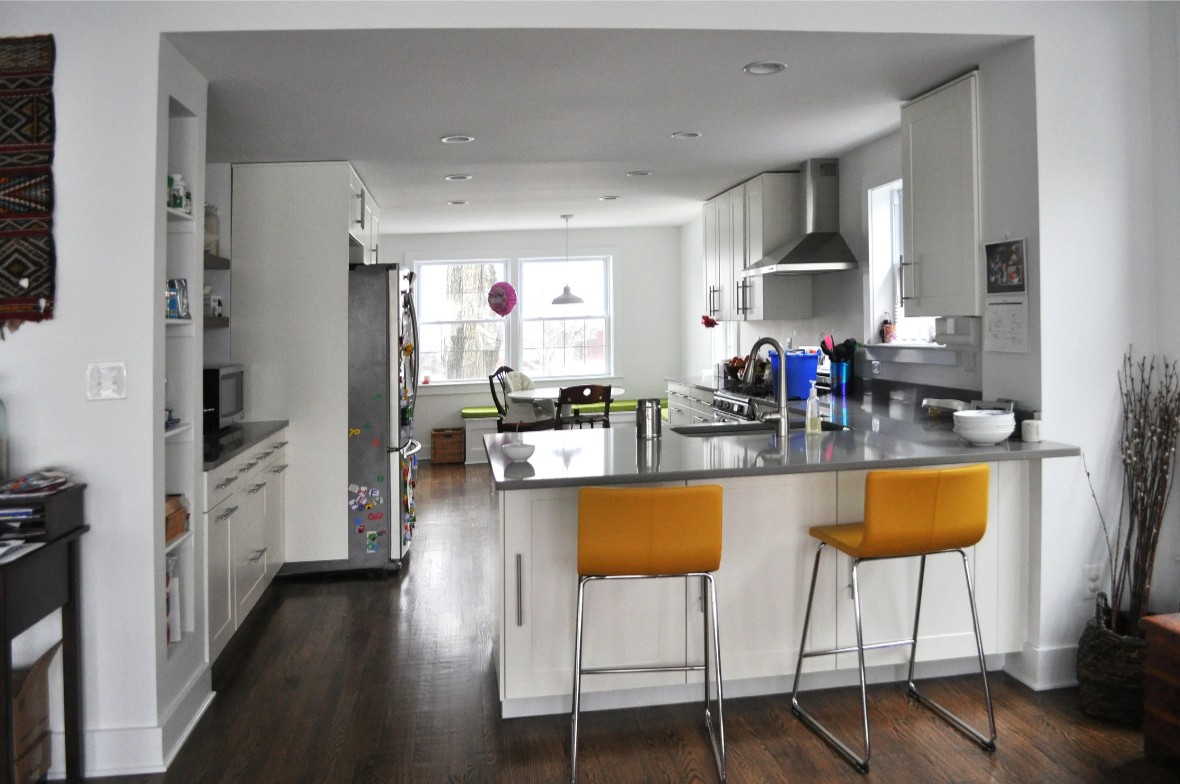 The renovated kitchen, expanded into the former dining room: white casework with gray quartz counters, two yellow stools at the counter peninsula in the foreground, a stainless hood over the cooktop, and a banquette under a pair of windows at the far end of the room