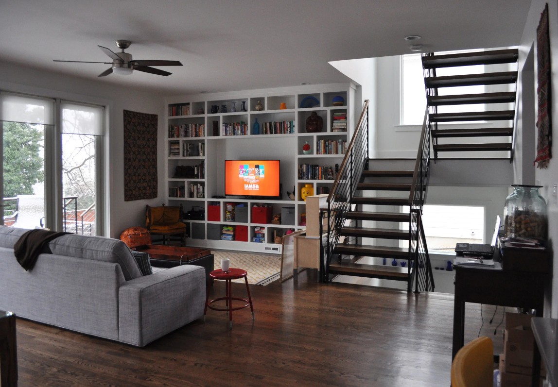 The same living room from the opposite side: a wall of built-in white bookshelves carries the back wall, a gray sofa sits in front, and an open metal-stringer stair with cable rails climbs to the master suite above