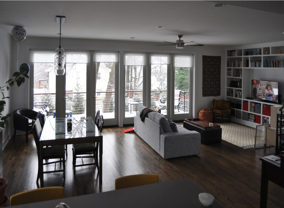 The living and dining wing of the addition looking toward the rear: a full-height glazed wall opens onto the deck and the snow-covered yard beyond, with the dining table at left and a gray sofa, ottoman, and built-in white bookshelves at right