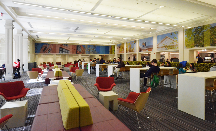 A second view of the commons from the opposite end of the room, the lounge zone of banquettes and red chairs in the foreground giving onto the counter-height dining zone and the servery line beyond, the photo-mural clerestory band running the length of the room