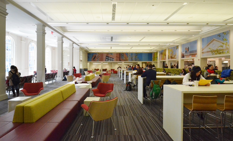 The dining commons looking down its length: tall arched windows and a colonnade along the left wall, an upholstered banquette and red lounge chairs in the foreground, counter-height dining tables running into the distance, and the photo-mural clerestory continuing above the servery line at right