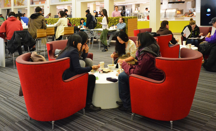 Four students sitting in a cluster of red tub chairs around a small round table, eating, with the busier room and the servery counter visible behind them