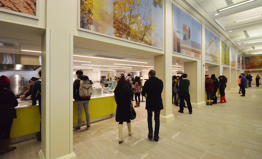 Students in line at the curved yellow servery counter, a worker preparing food behind the line, the stainless hood and kitchen visible above and behind