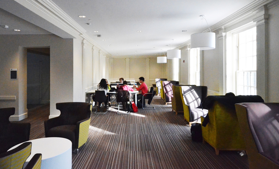 The upstairs study room with tall windows, high-backed lounge chairs in mustard and dark upholstery, and a worktable in the middle of the room where three students are working together