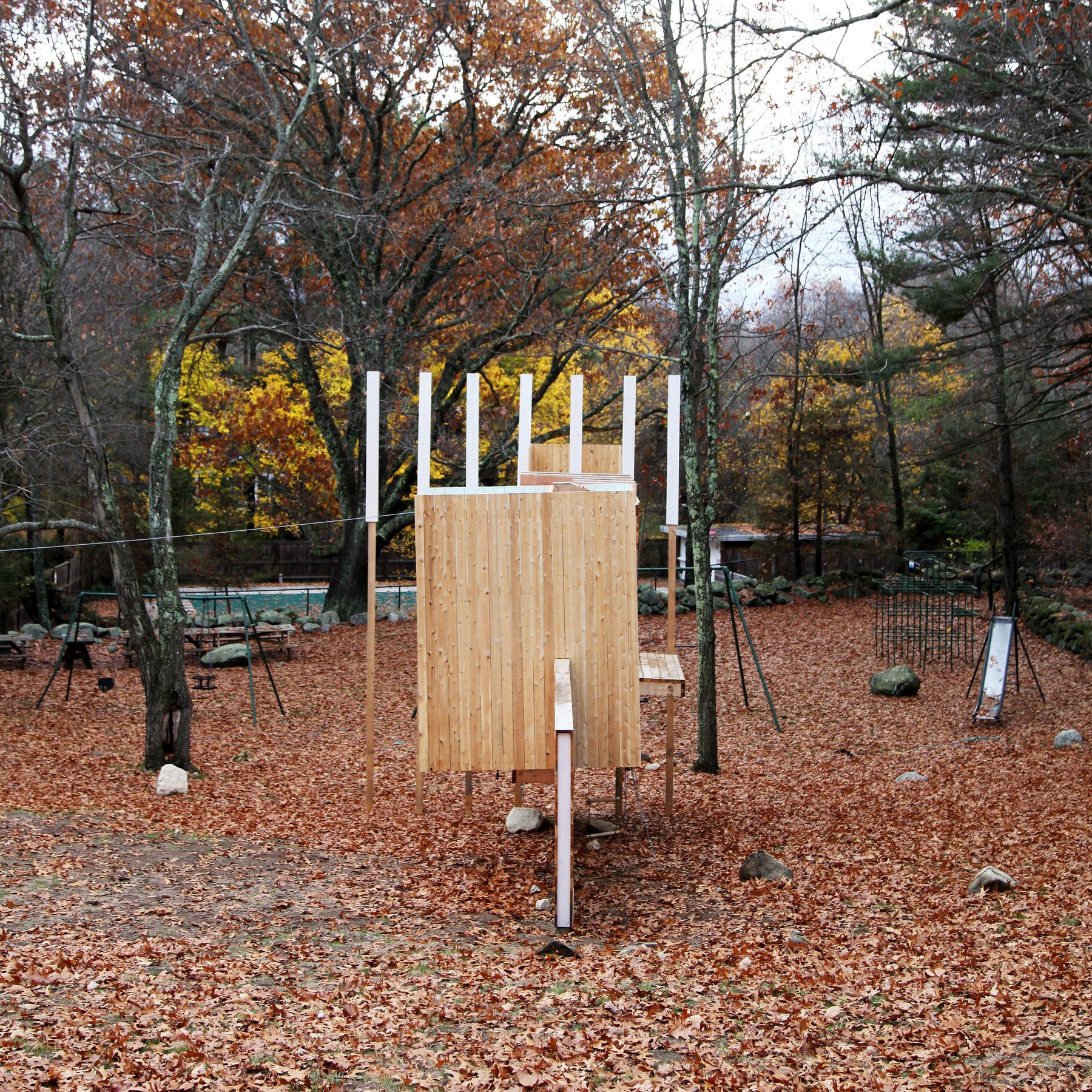 The structure seen from behind, with the existing slide, swings, and sandbox visible in the surrounding clearing