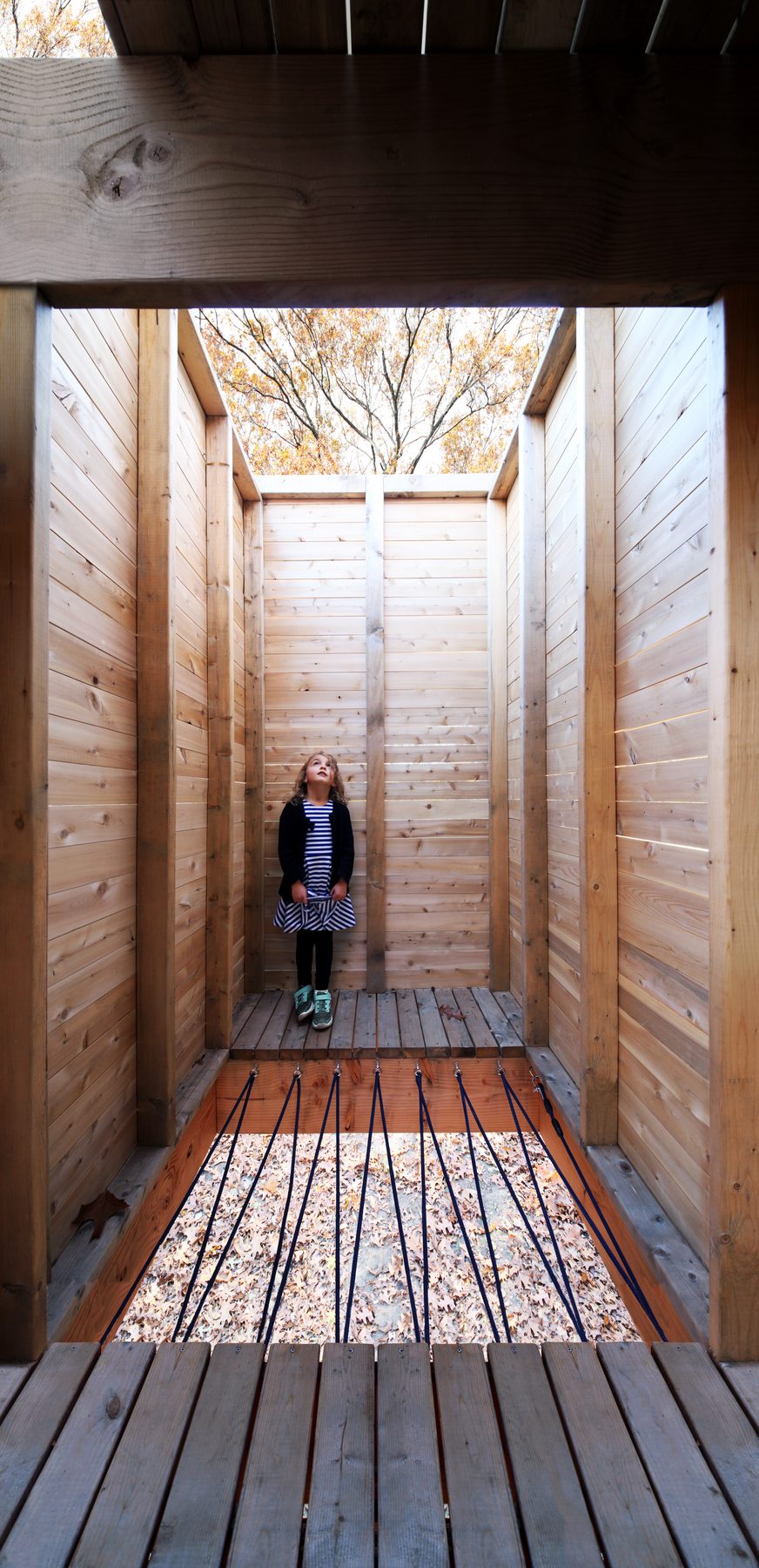 Looking through one of the wood rooms with a child standing in striped shirt, looking up toward the open sky, a rope-bridge floor in the foreground