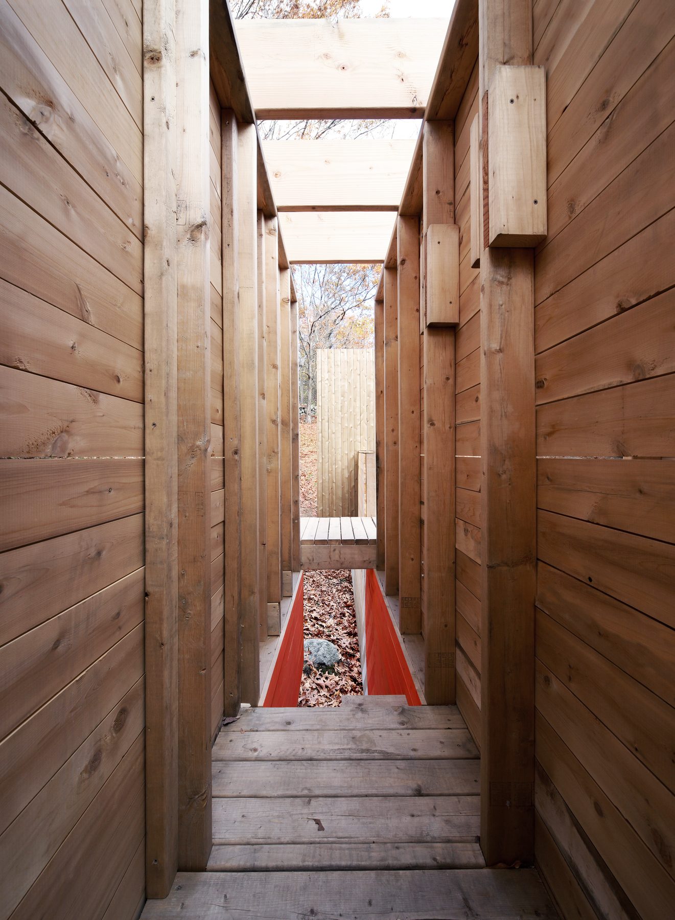 A narrow slot interior looking toward an orange-painted slide-channel below and a doorway opening to the landscape beyond