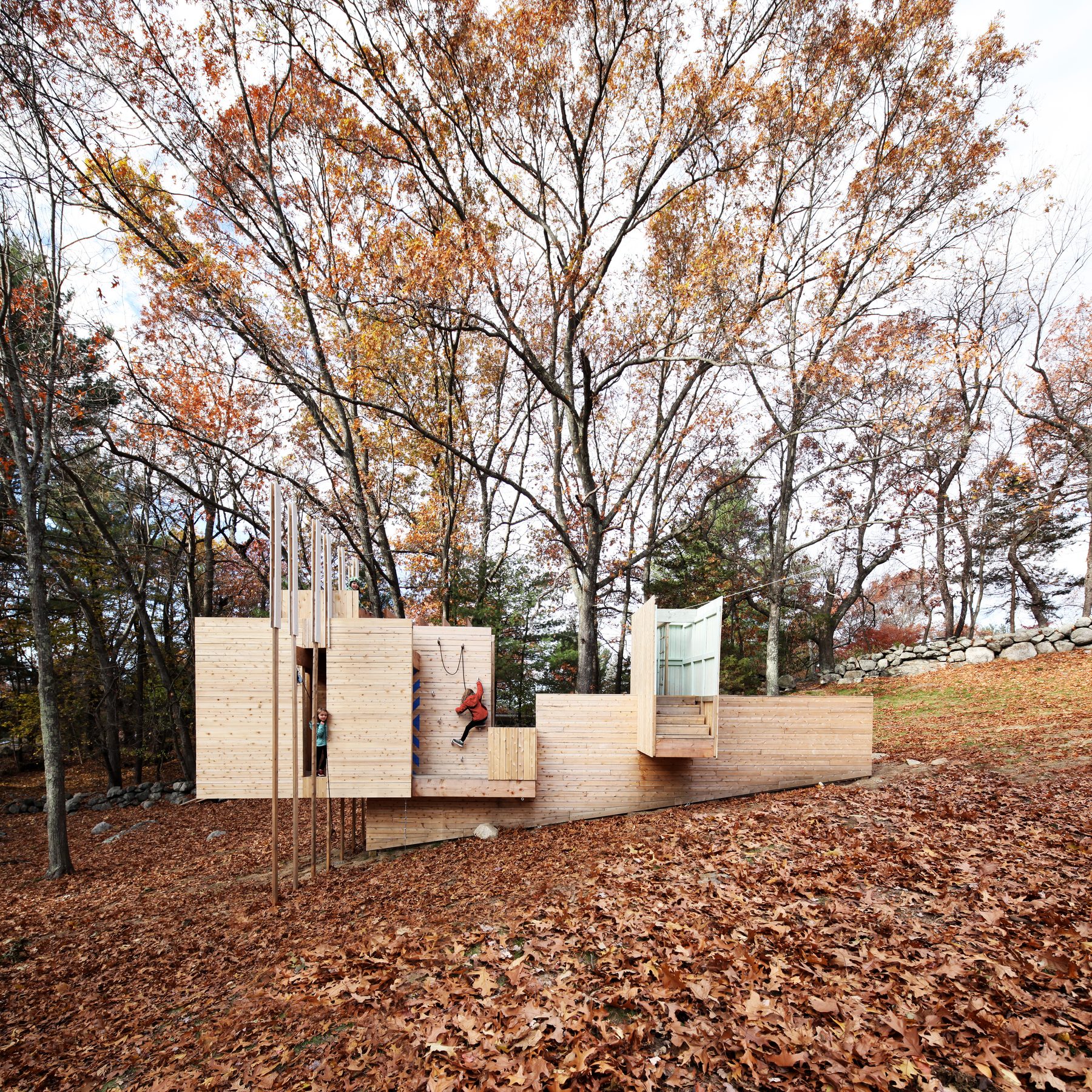 The play structure in autumn, set in a clearing of fallen leaves with two children visible — one mid-climb on the rope, one in the doorway