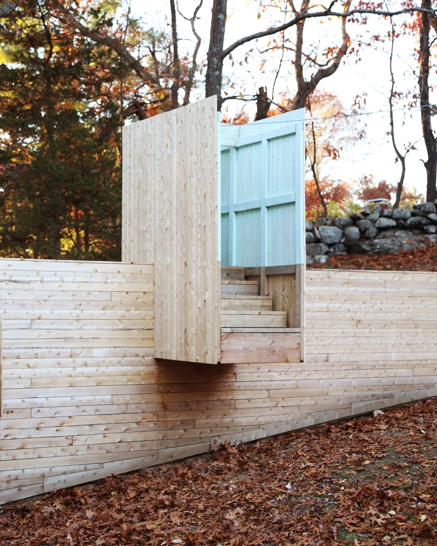 A robin&rsquo;s-egg blue door standing ajar above the wood wall, with stairs visible beyond and a stone wall in the autumn woods behind