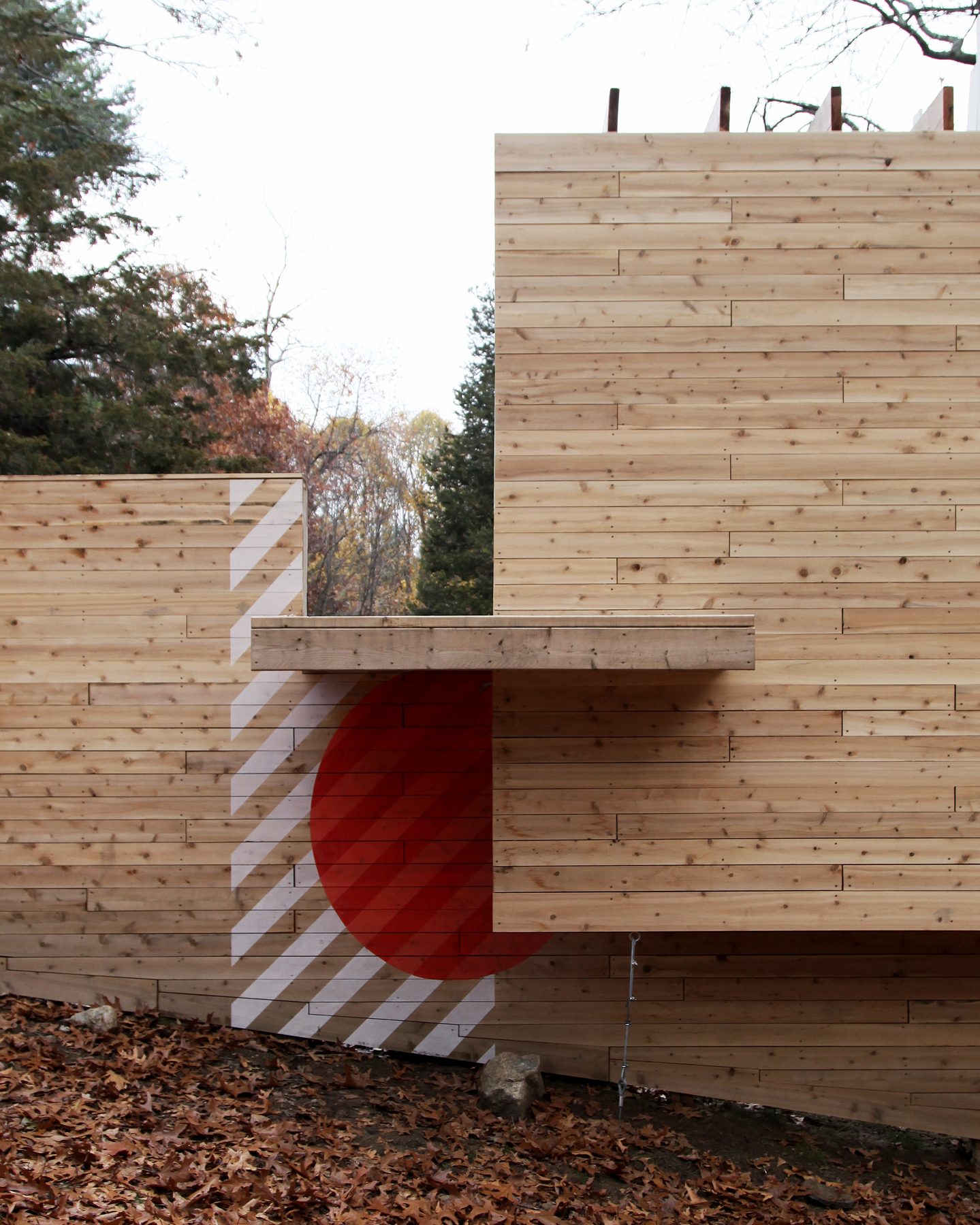A cantilevered shelf above a painted red circle and white stripes, with a slot through to the woods beyond