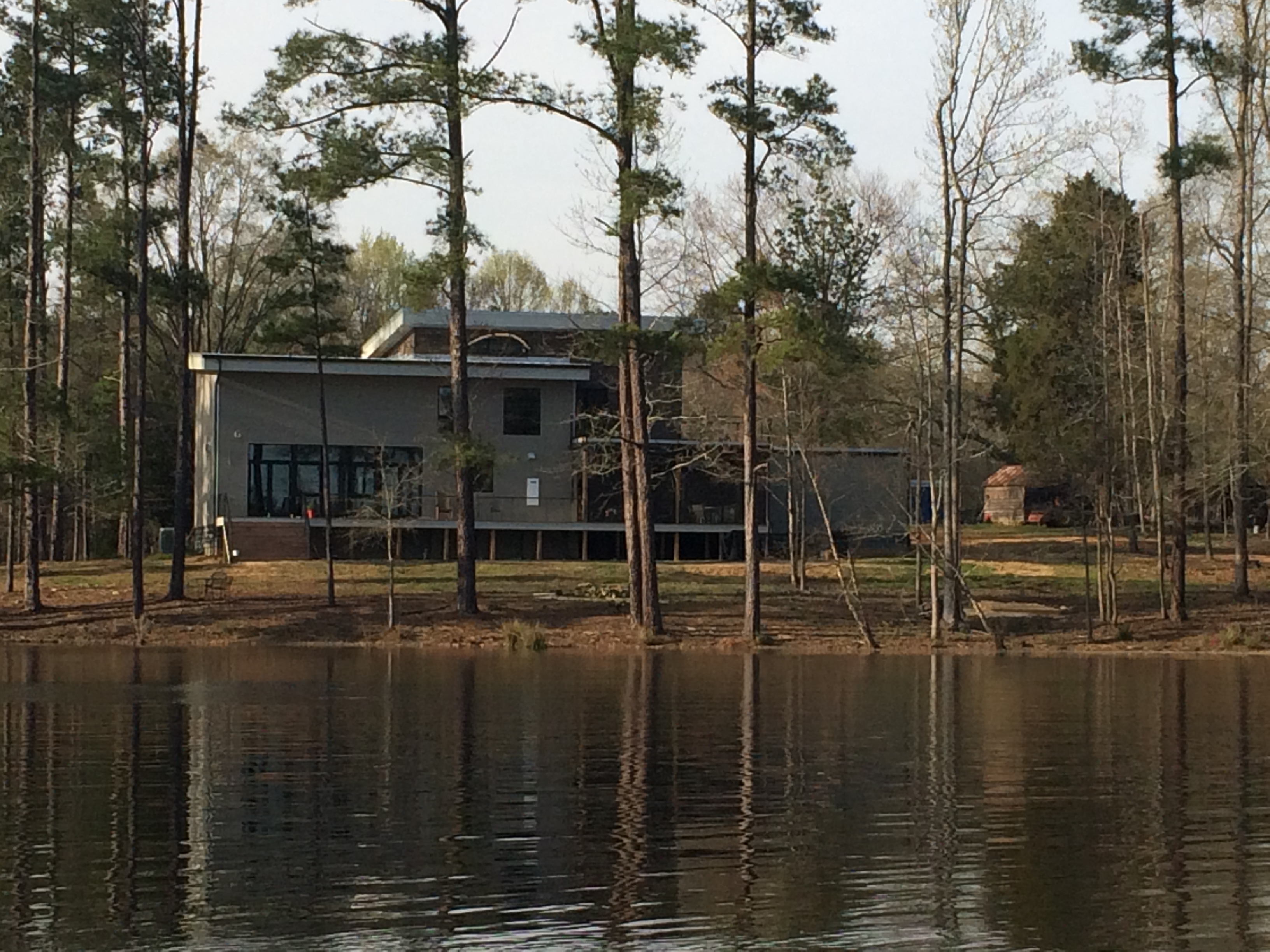 The house seen from across the lake, sitting on its small hill among tall pines, lifted lightly on piers above the slope, with a reflection in the still water in the foreground