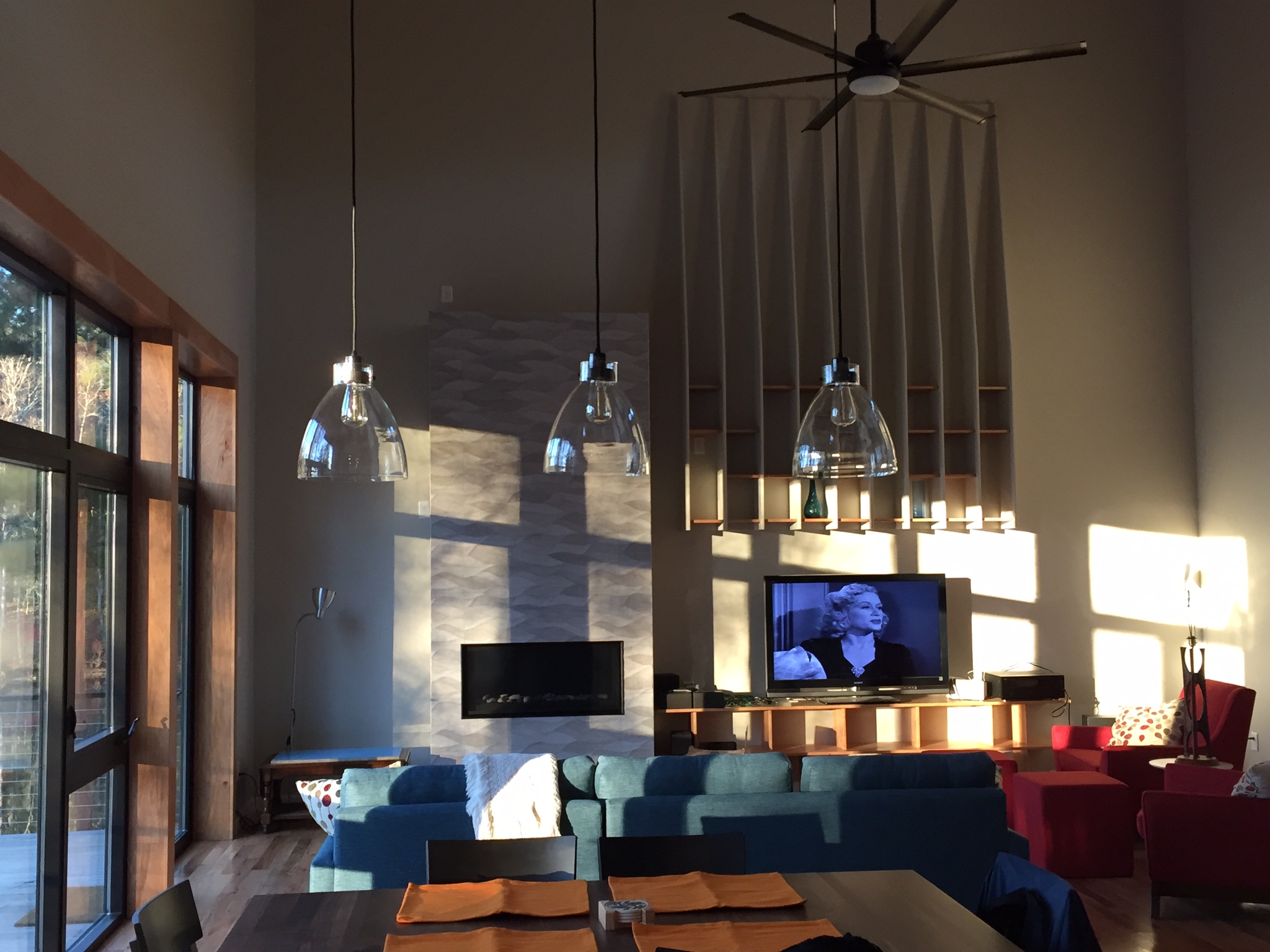 The living room at evening, looking toward the textured stone fireplace, the pendants hanging the full height of the double-height room, and the tall sheer drapery beside the ceiling fan