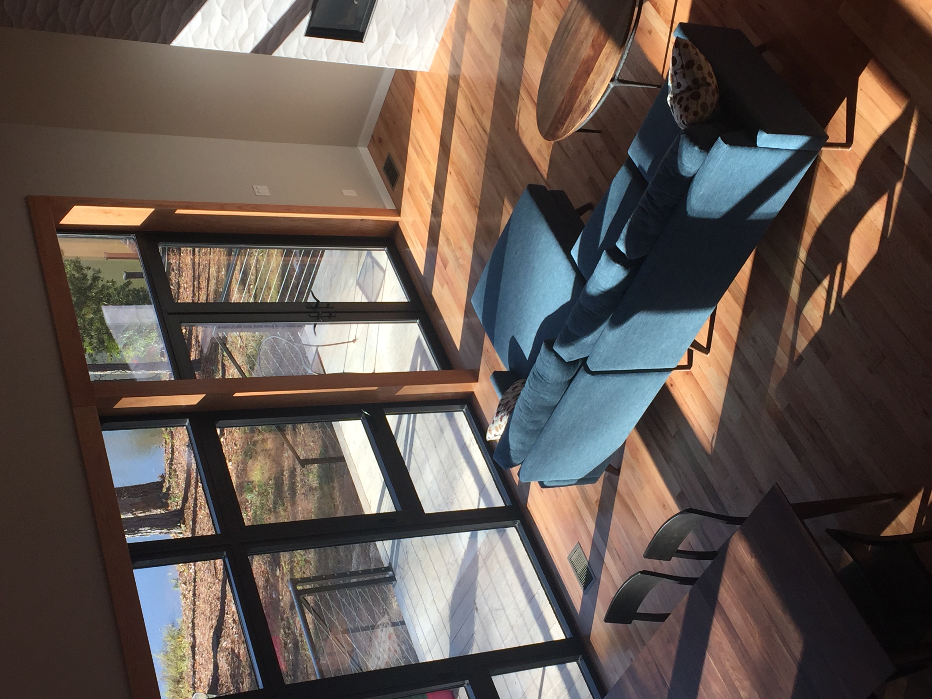 View down from the upper loft into the double-height living room, with the blue sectional sofa, the dining chairs, and the tall glazing on the lake side throwing strong winter light across the floor