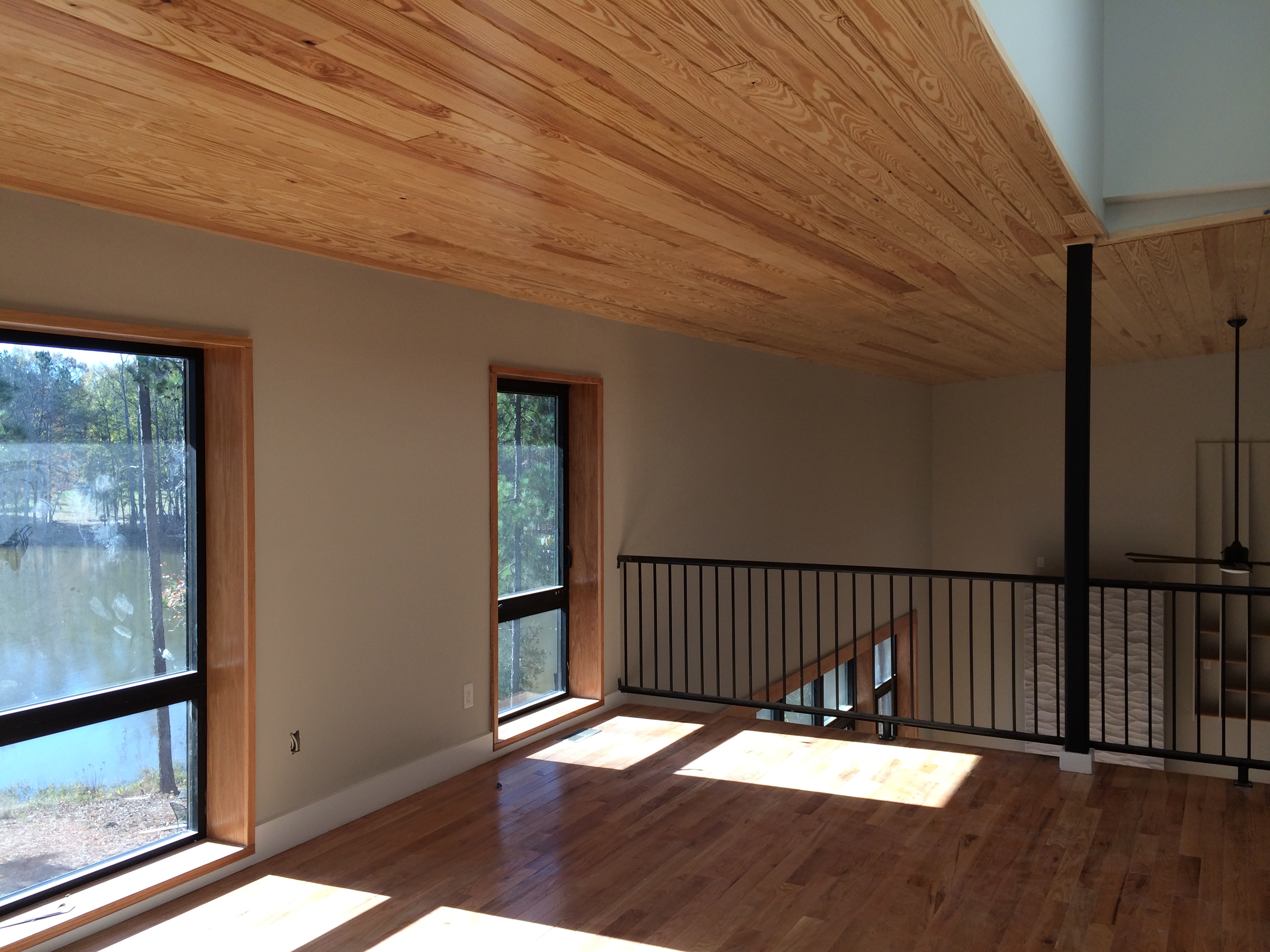 The upstairs loft with the wood ceiling raking up toward the ridge, two tall windows framing the lake and the pines, and the open stair railing visible to the right