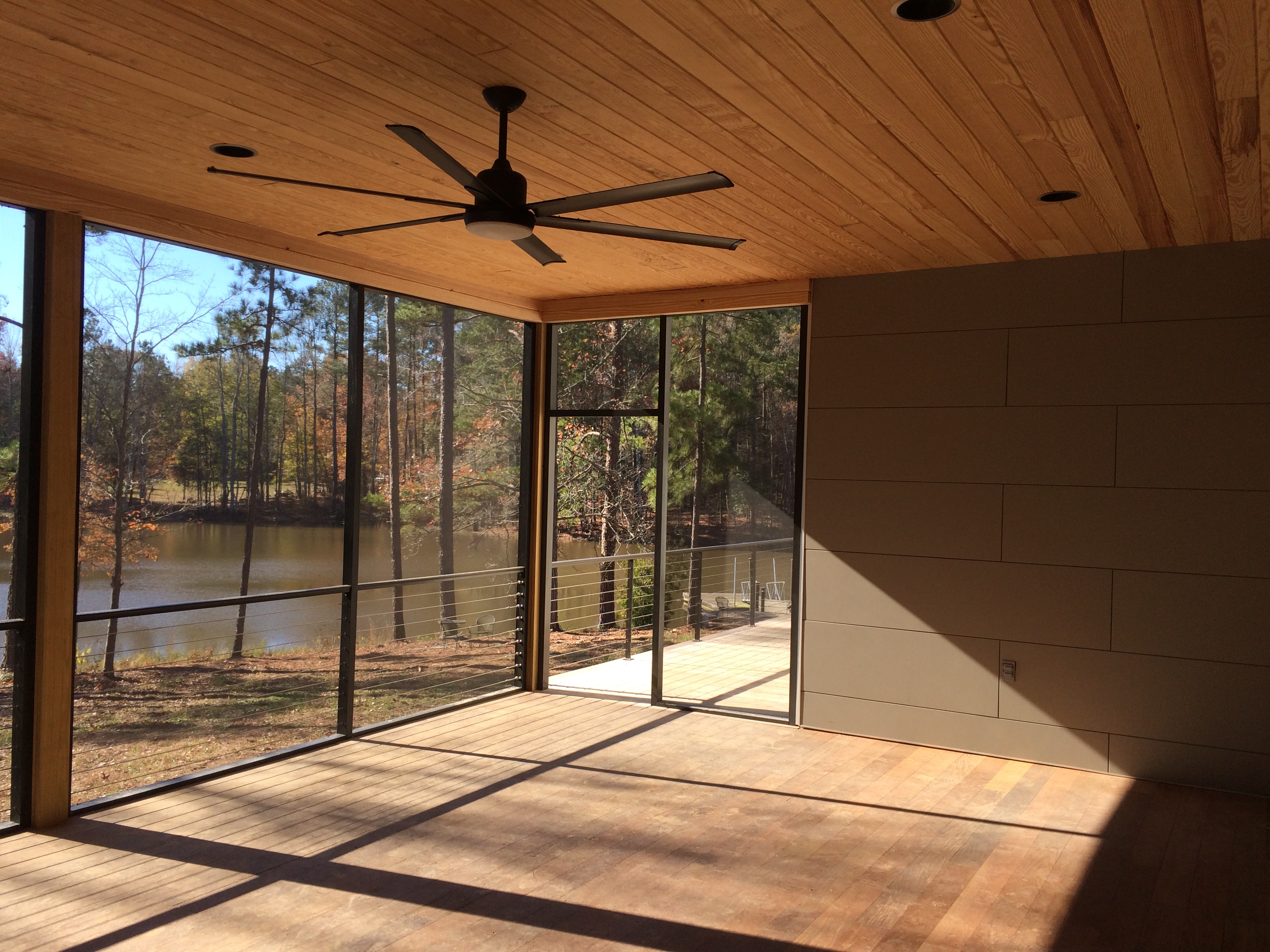 The screened porch on the lake side, the wood ceiling continuing from inside, a large ceiling fan overhead, and the pines and the water visible through the full-height screens