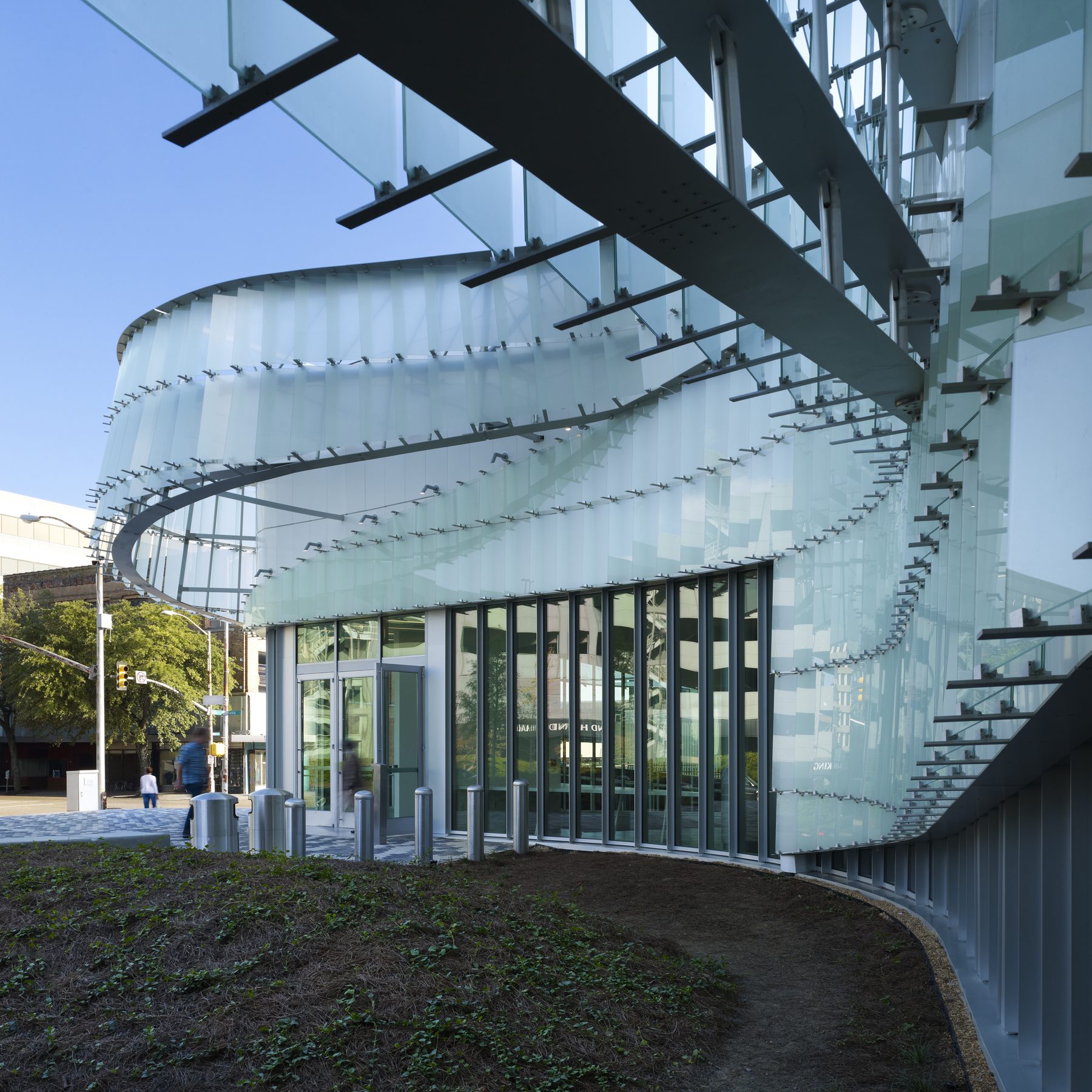 The pavilion at the street corner, the steel framing visible beneath the curving glass-shingle skin