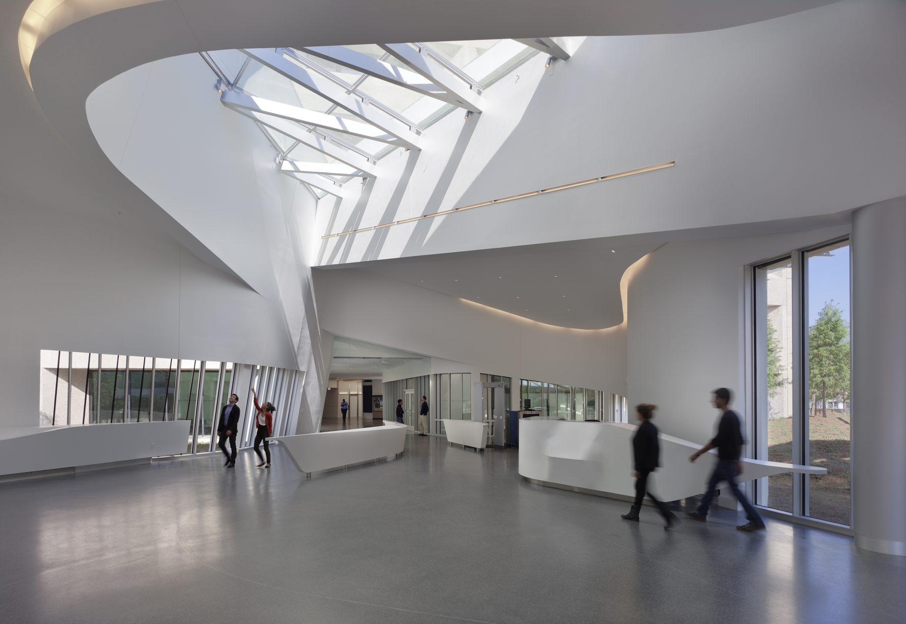 The skylit lobby with the spiral geometry resolving overhead into faceted white planes, people moving through the screening floor