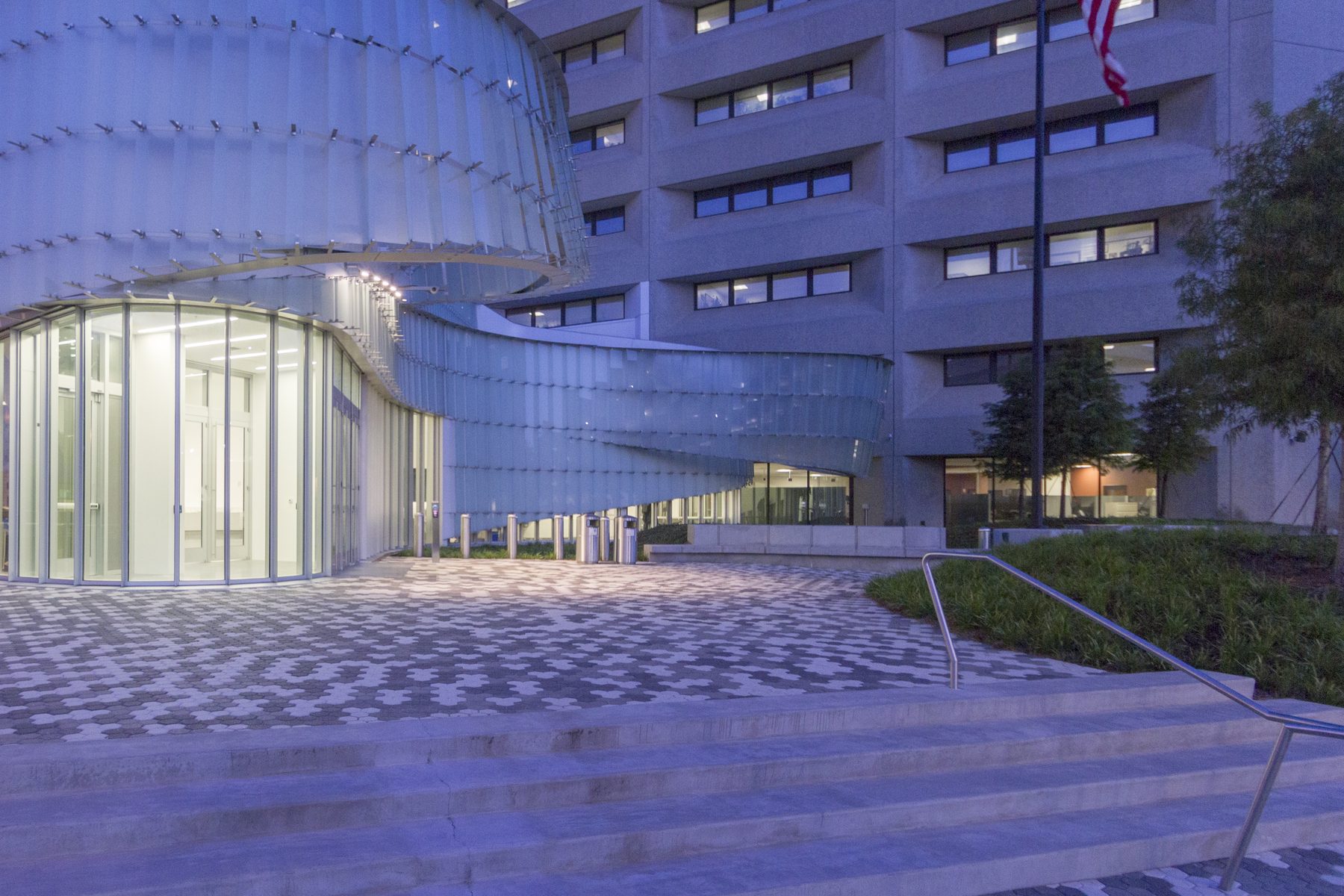 The pavilion at dusk read as a lantern from the entry plaza, the lobby visible through the translucent skin and the curving glass skin sweeping around toward the tower