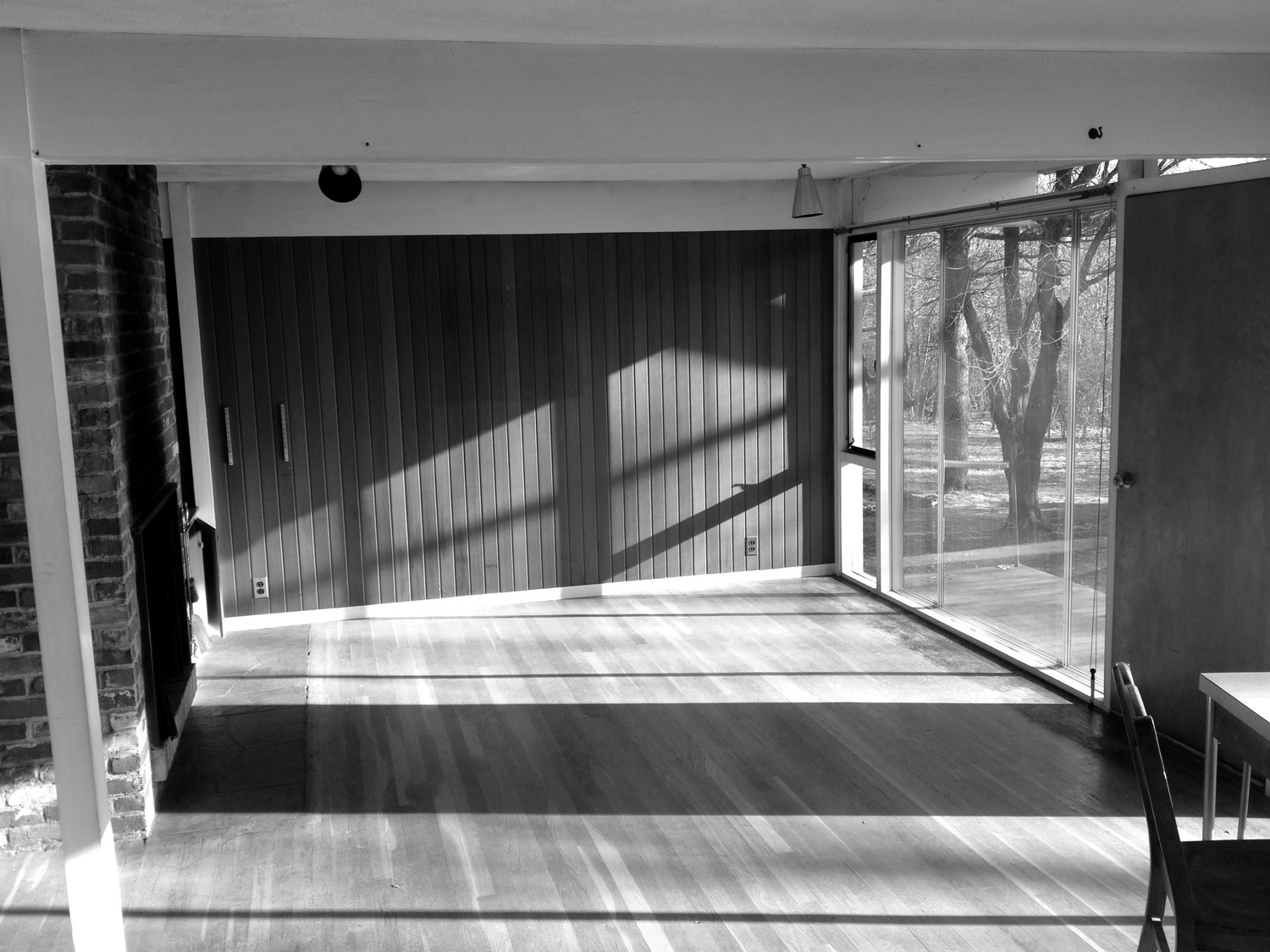 The original living room photographed before any work began: empty floor, mid-century vertical wood paneling on the far wall, the edge of the brick chimney at left, a slim ceiling beam, and floor-to-ceiling sliding glass at right giving onto bare winter trees