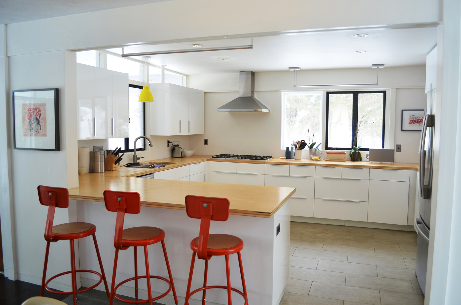 The renovated kitchen seen from the new opening: a U-shaped island with butcher-block counter and three red bar stools, white casework along two walls, a stainless hood over the cooktop, and tall windows above the counters looking out to bare winter trees