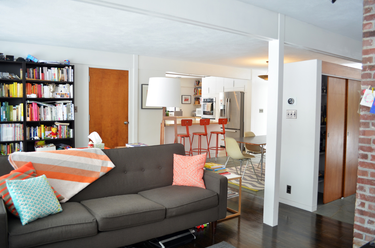 View from the living room across the back of a gray sofa, looking through the new opening in the central wall to the dining table and the kitchen beyond, with red counter stools at the bar and the orange book spines on the bookshelf at left