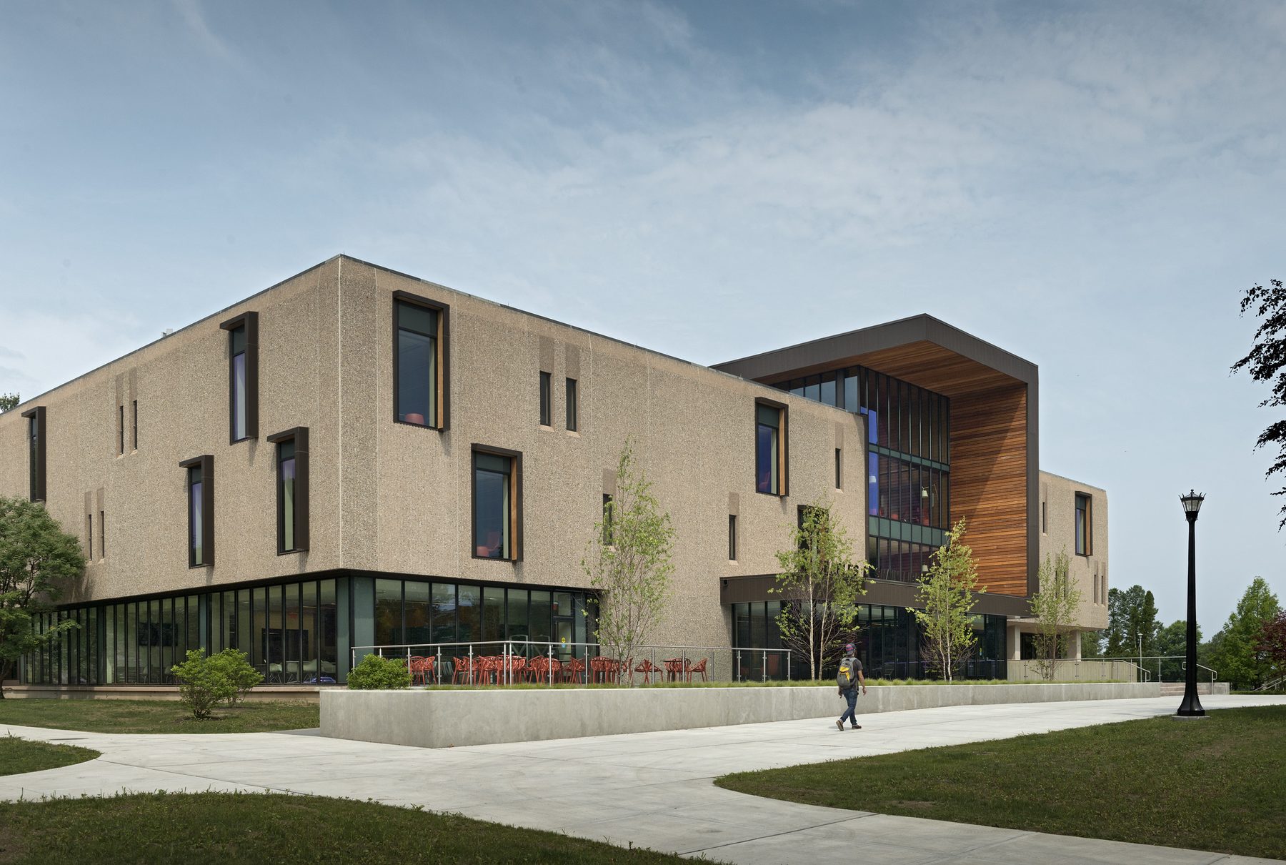 The renovated library in daylight, three-quarter view, with a single student walking past the cafe terrace and red chairs visible against the textured precast wall