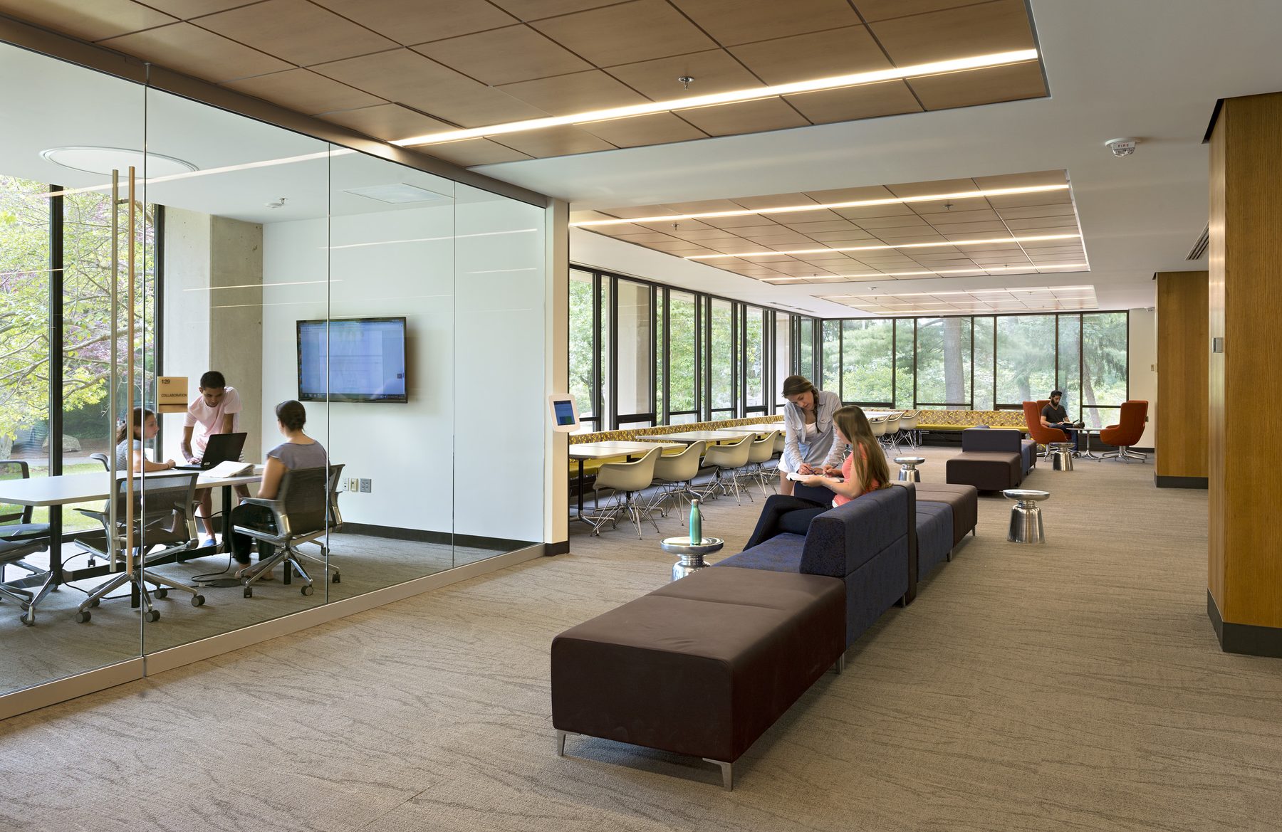 A study floor with a glass-walled collaboration room on the left and the new continuous window band along the back wall, with students reading and working at the tables