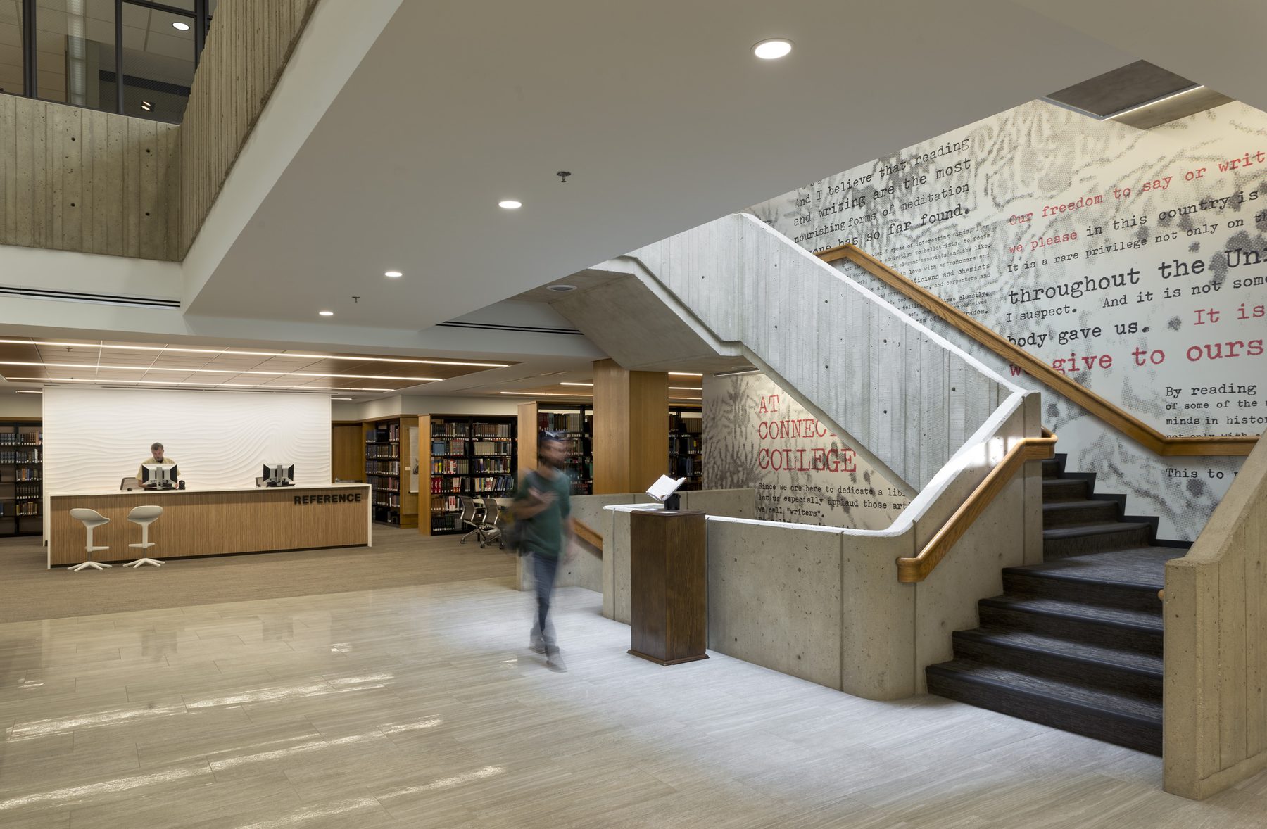 The central atrium of the library, with the board-formed concrete stair on the right rising past the typewriter-text mural drawn from Vonnegut's dedication speech, the reference desk visible at left, and a blurred student walking through
