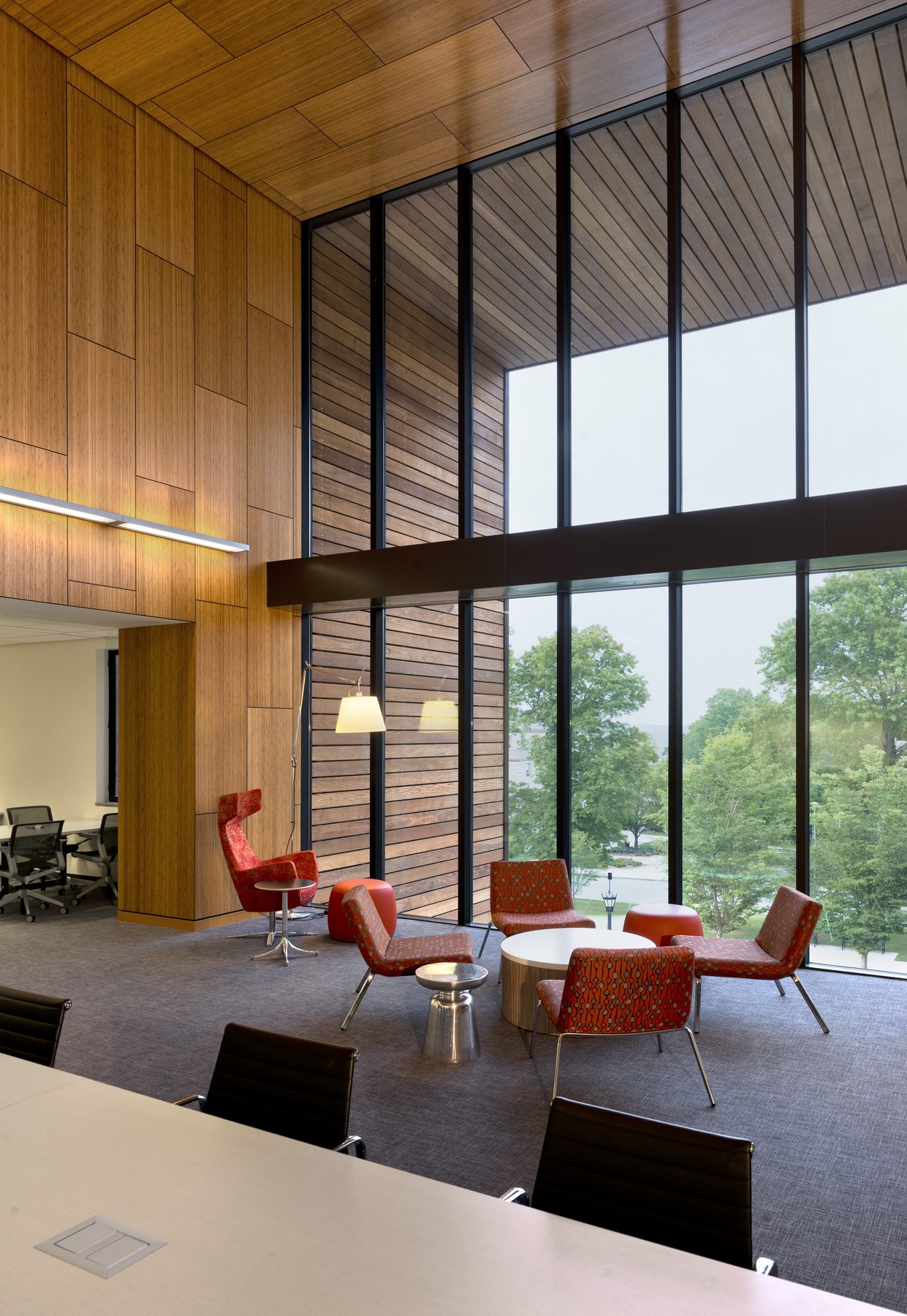 The same wood-lined volume seen from inside, looking out through full-height glazing to the trees and campus beyond, with red chairs in the foreground and bamboo paneling rising the full two stories
