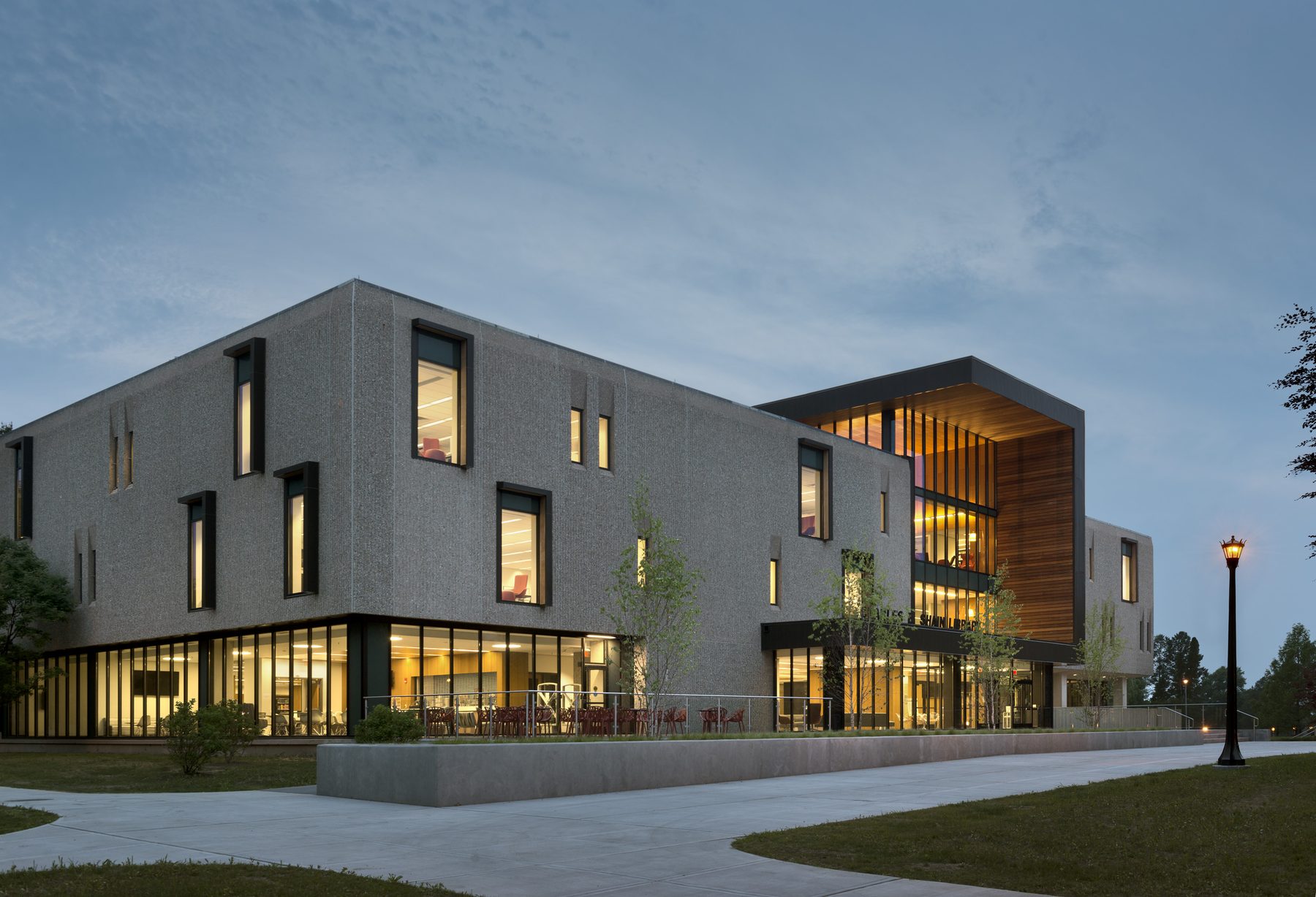 The renovated library at twilight, three-quarter view: the original precast volume in shadow with interior light glowing through the slit windows, and the wood-lined entry incision lit from within