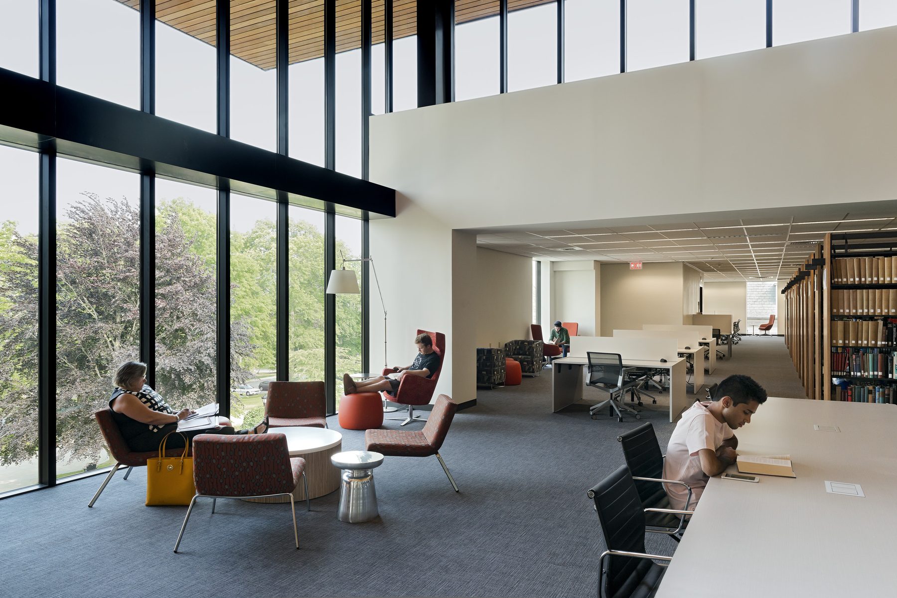 A reading lounge inside the wood-lined volume with full-height glazing on the left, students reading at the windows, and the book stacks visible at right