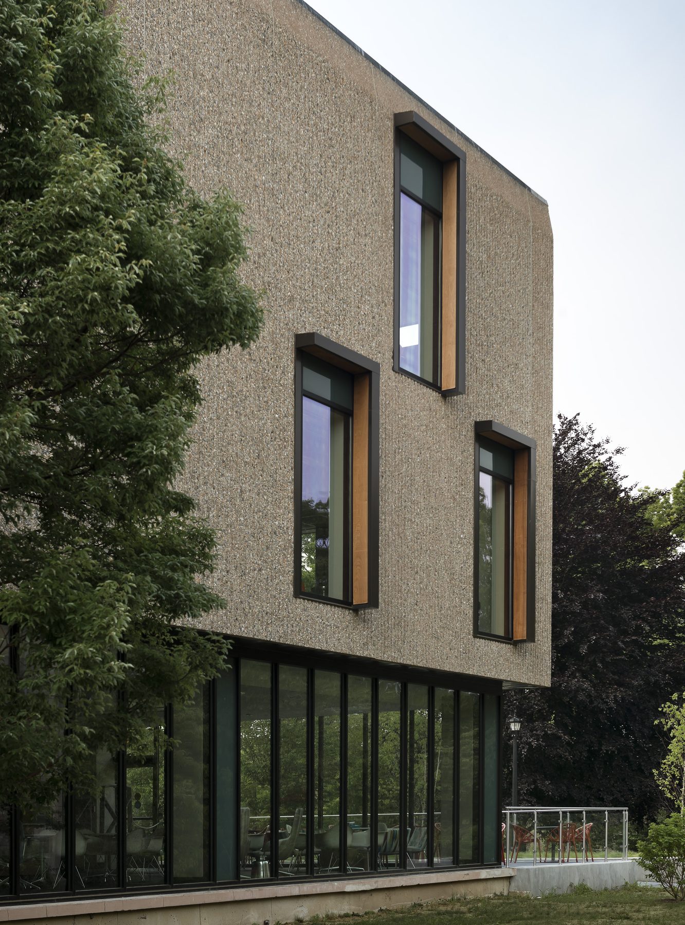 Three new vertical window openings cut into the textured precast wall, each lined with warm wood reveals, with the cafe glazing visible below where part of the moat was filled