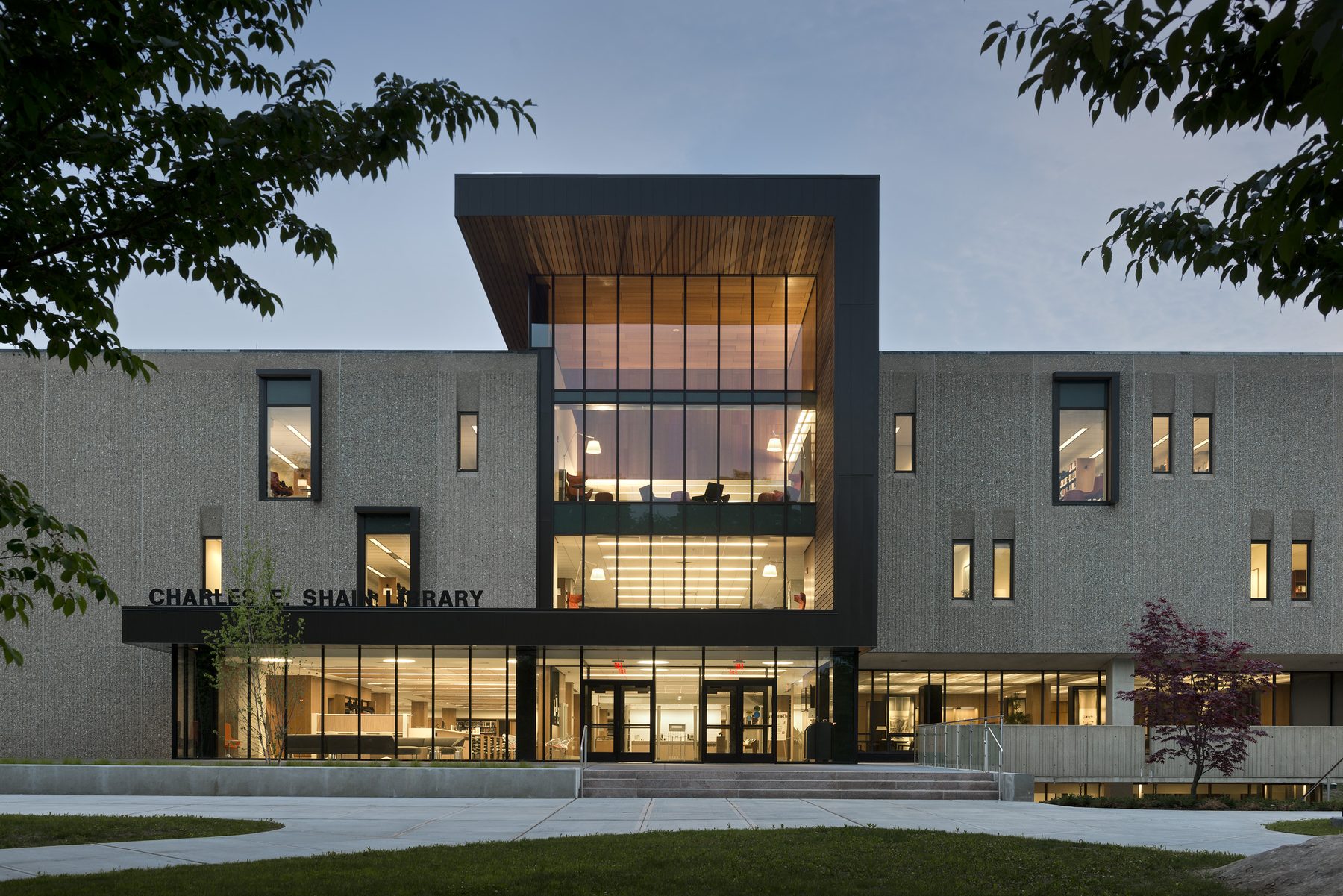 The renovated Shain Library at twilight, head-on view: original textured precast wall on either side of a glowing wood-lined volume that has been cut through the front facade as the new entry